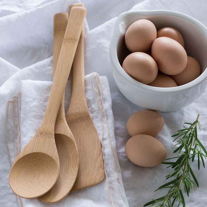 Wooden spoons on a white cloth with a bowl of eggs and rosemary.
