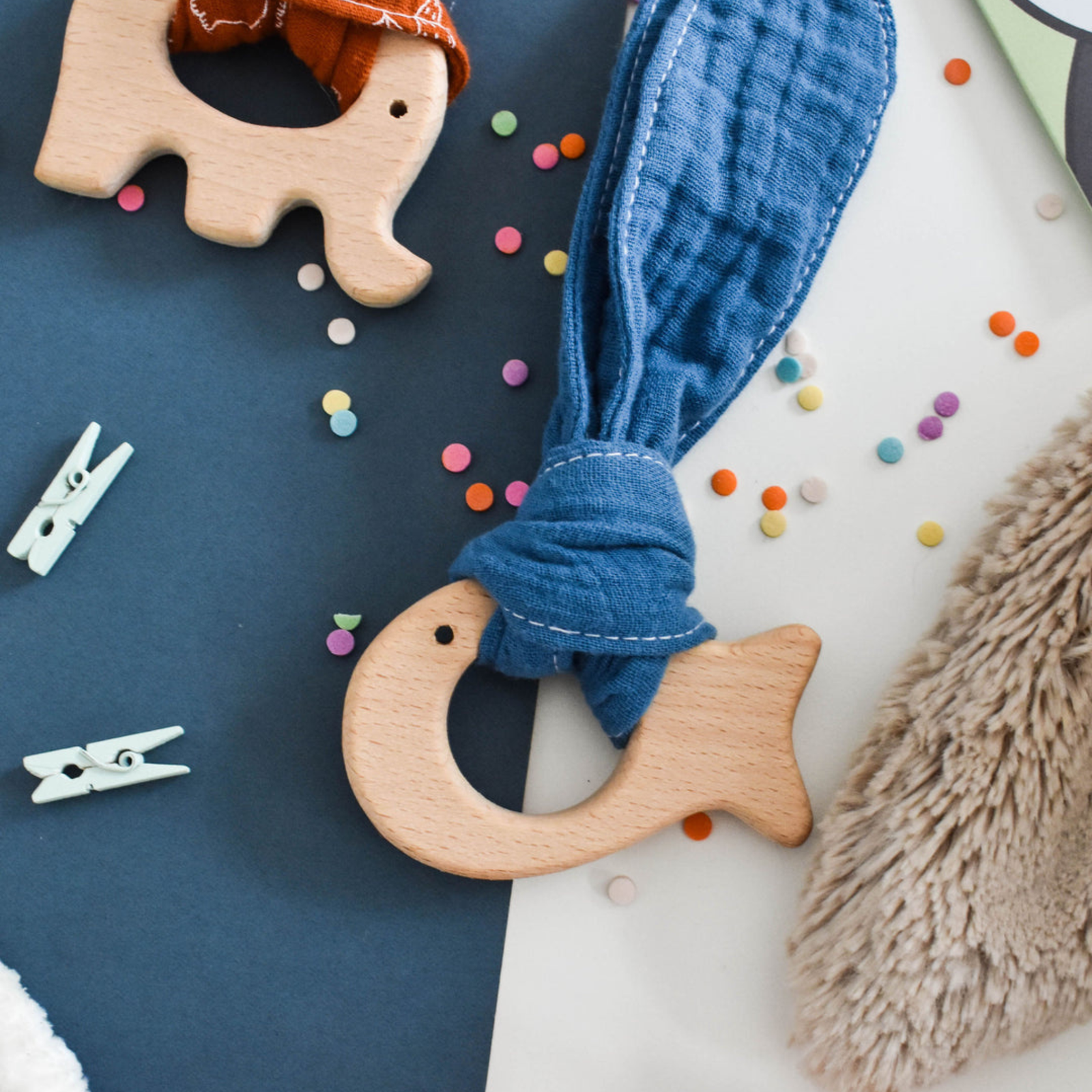 Wooden fish teether with blue fabric, colorful confetti, and white clothespins on a dark surface.