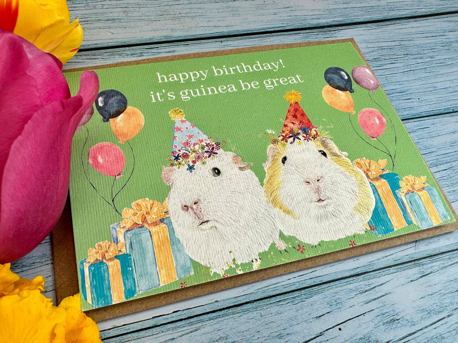 Birthday card with two guinea pigs wearing party hats on a wooden surface with flowers.
