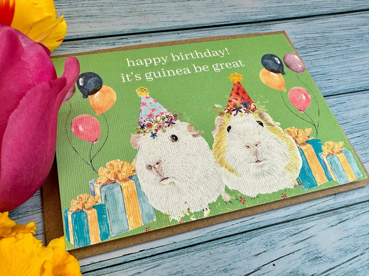 Birthday card with two guinea pigs wearing party hats on a wooden surface with flowers.