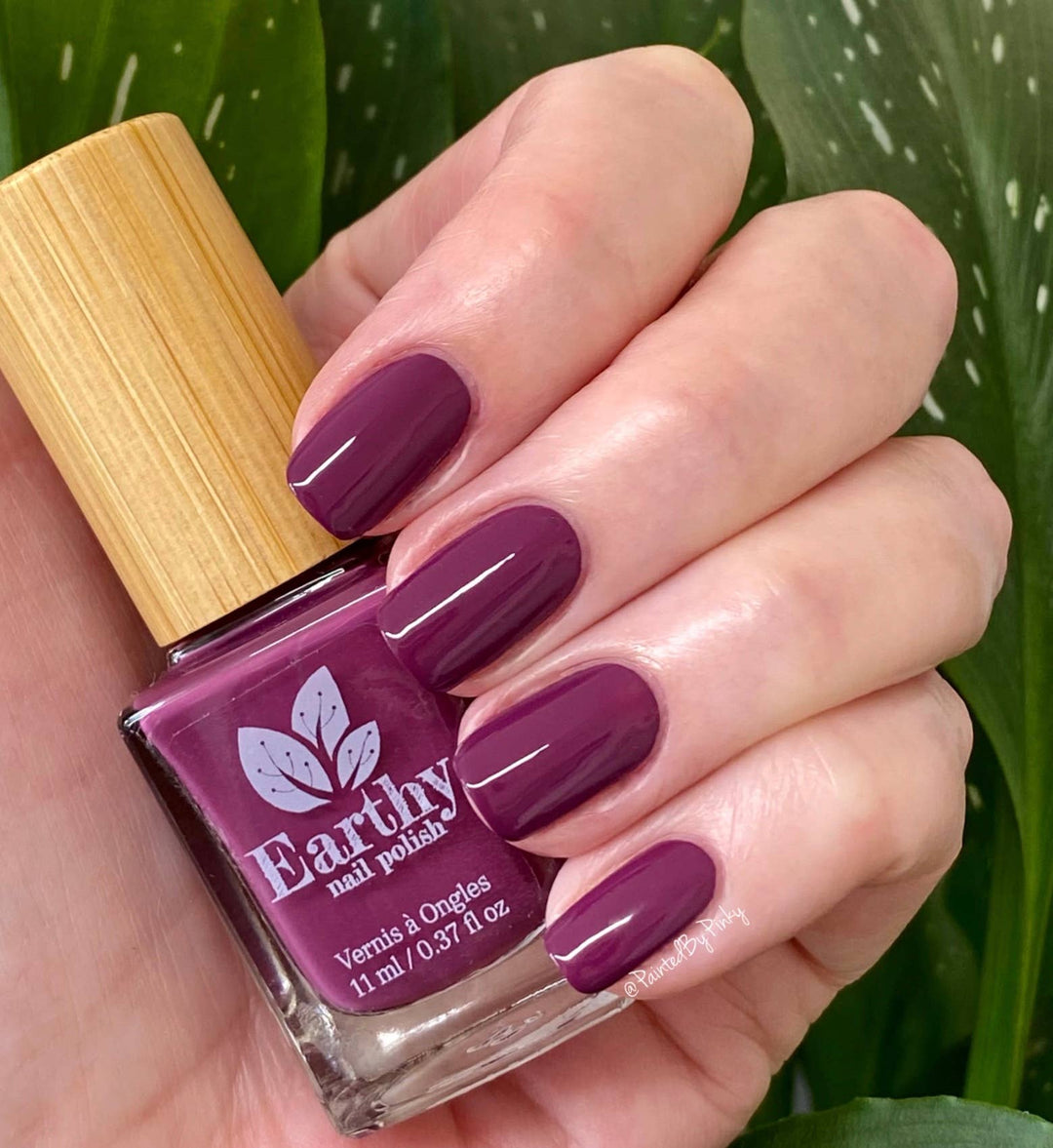 Hand with purple nail polish holding an Earthly nail polish bottle against a green leafy background