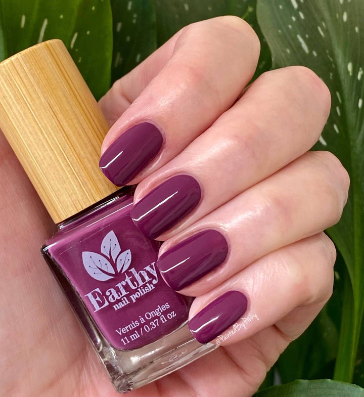 Hand with purple nail polish holding an Earthly nail polish bottle against a green leafy background