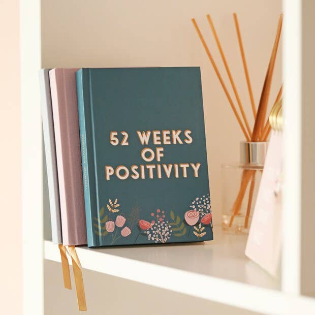 Write down your thoughts, feelings and reflections on each day of the year, this diary focuses on the positive. The pages are encased in a teal cover adorned with illustrations of pink flowers and botanical details. The front is finished with the wording '52 weeks of positivity' in a bold font.