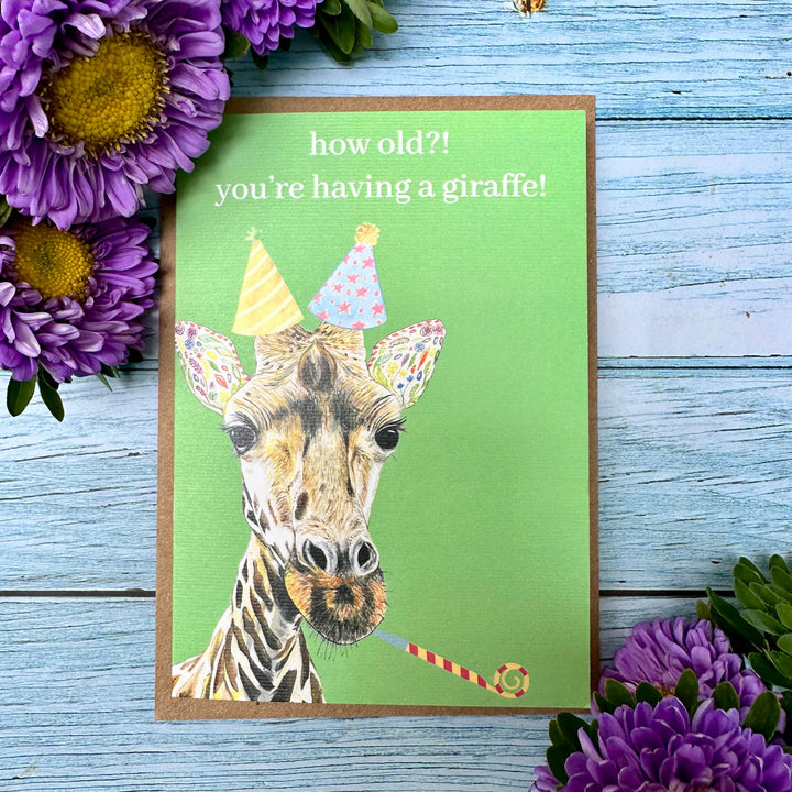 Greeting card with a giraffe illustration and birthday-themed text, surrounded by purple flowers on a wooden surface.