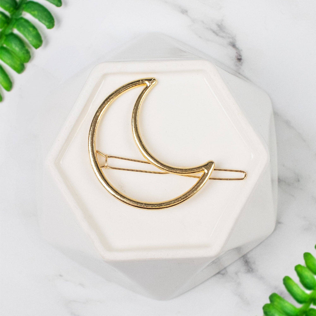Gold crescent moon hair clip on a white hexagonal stand with green leaves in the background