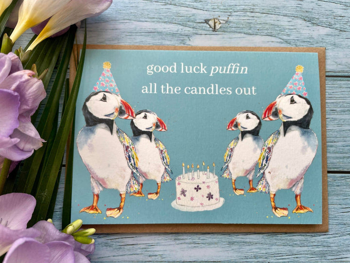 A greeting card with an illustration of puffins wearing party hats and a cake with the words 'good luck puffin all the candles out'. The card is placed on a wooden surface with purple flowers in the background.