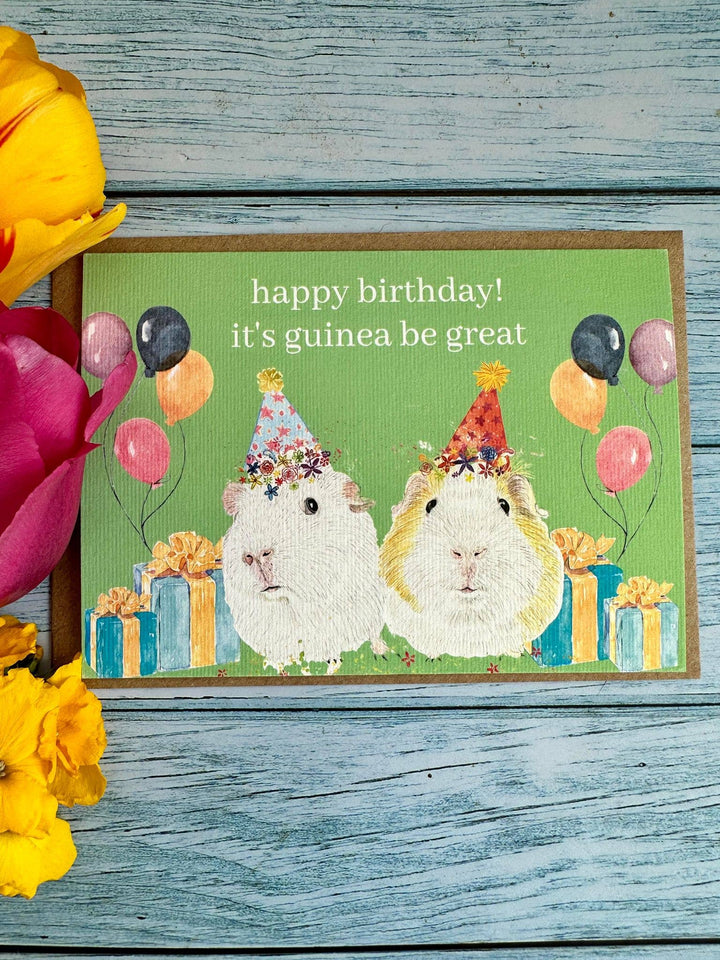 Birthday card with two guinea pigs and balloons on a wooden surface with flowers.