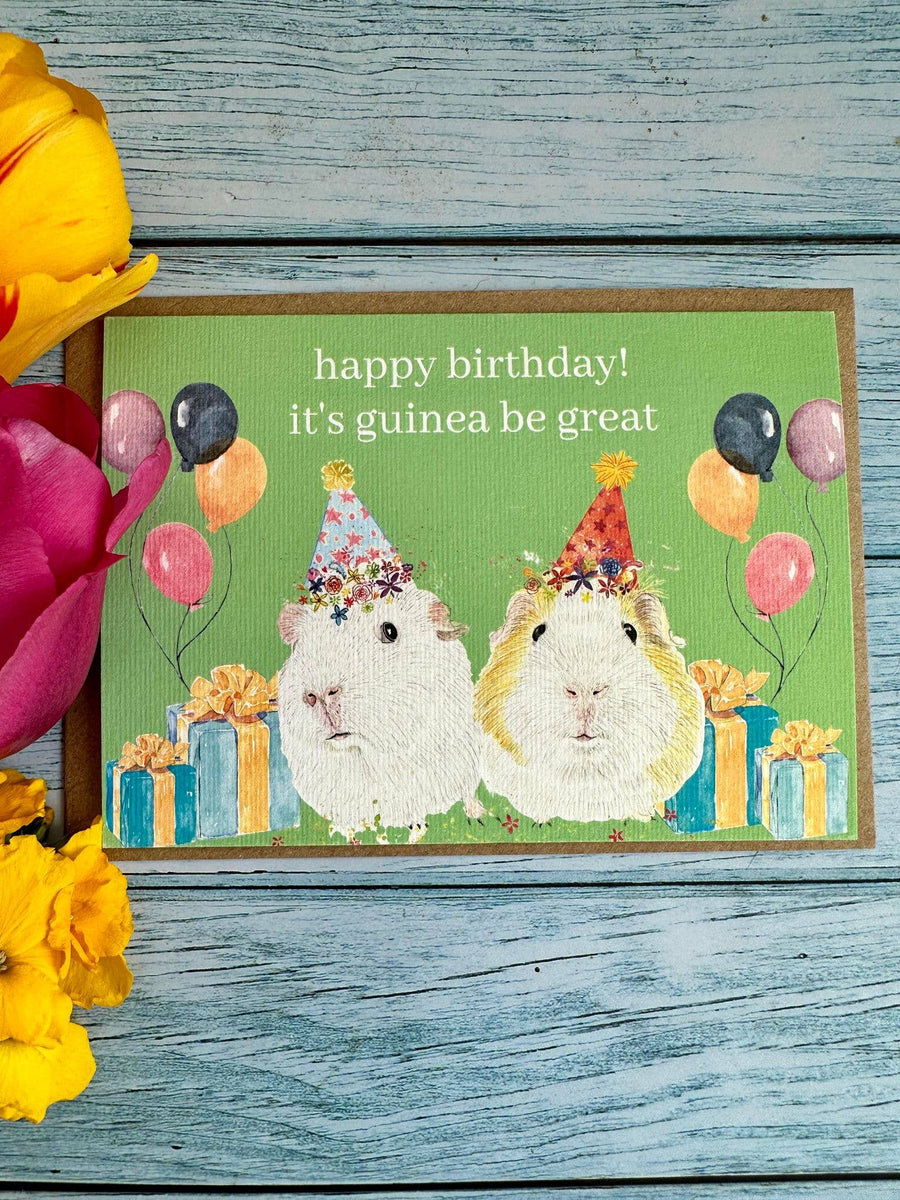 Birthday card with two guinea pigs and balloons on a wooden surface with flowers.
