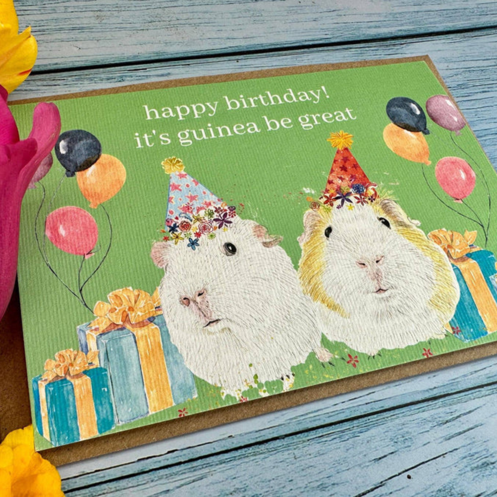 Birthday card with two guinea pigs wearing party hats and balloons on a wooden surface.