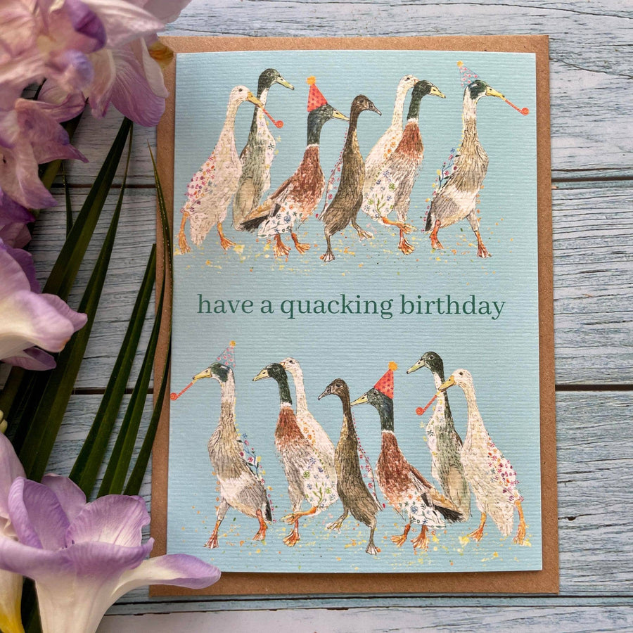 A greeting card with an illustration of ducks wearing party hats, with the text 'have a quacking birthday' on it. The card is placed next to some purple flowers.
