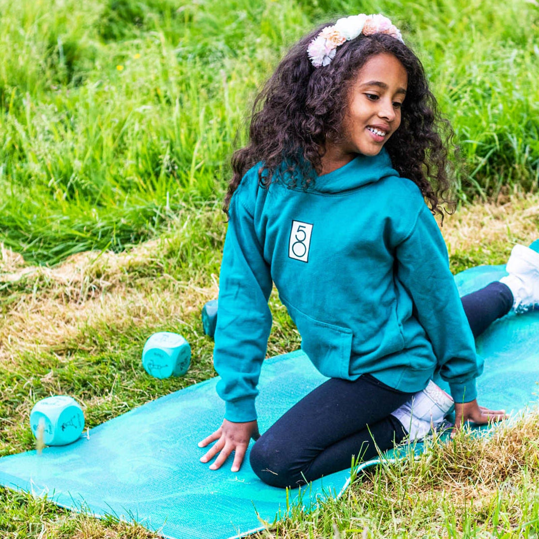 Young girl in a teal hoodie in a yoga pose from the dice on a blue mat outdoors on grass