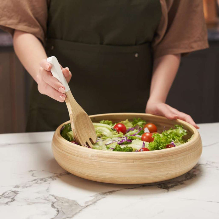 Person preparing a salad in a wooden bowl with a wooden spoon on a marble countertop.