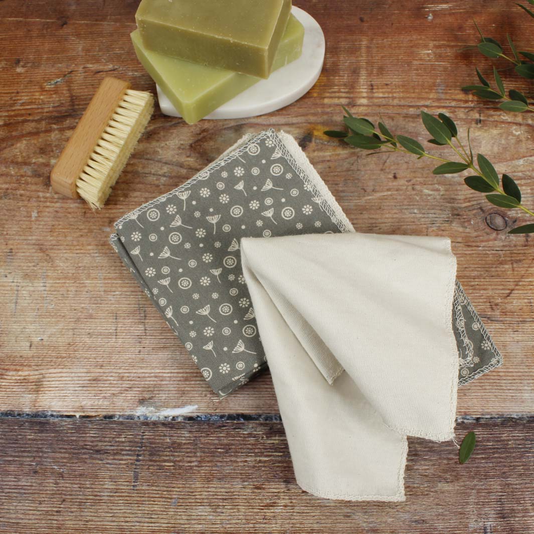 Two reusable cloths grey floral on a wooden surface with a scrub brush and soap bars.