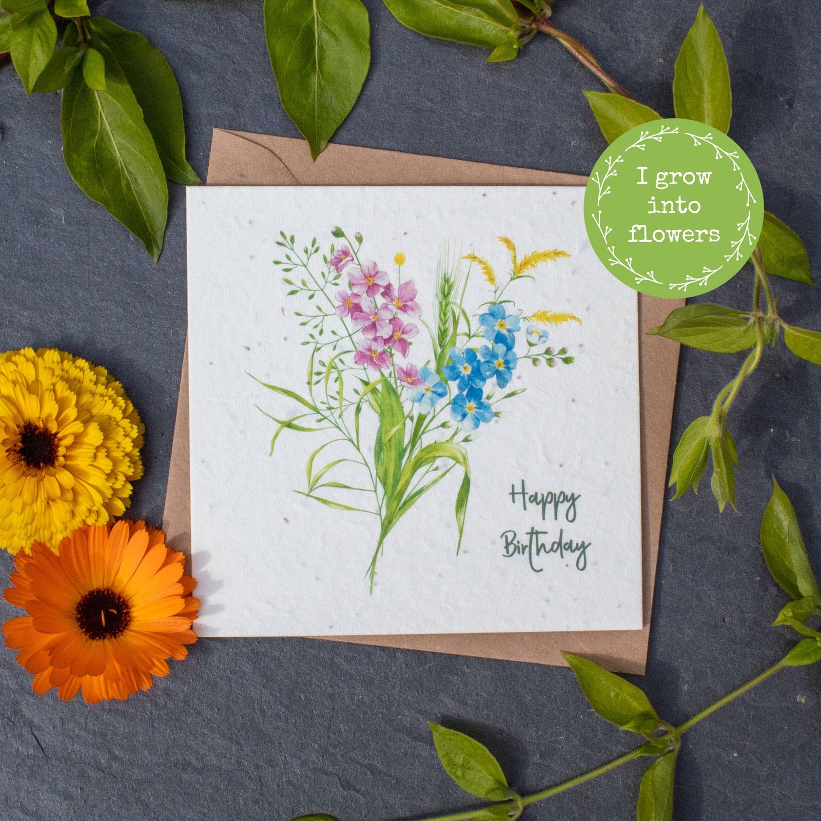Greeting card with floral design and 'Happy Birthday' text, surrounded by real flowers and leaves on a dark surface.