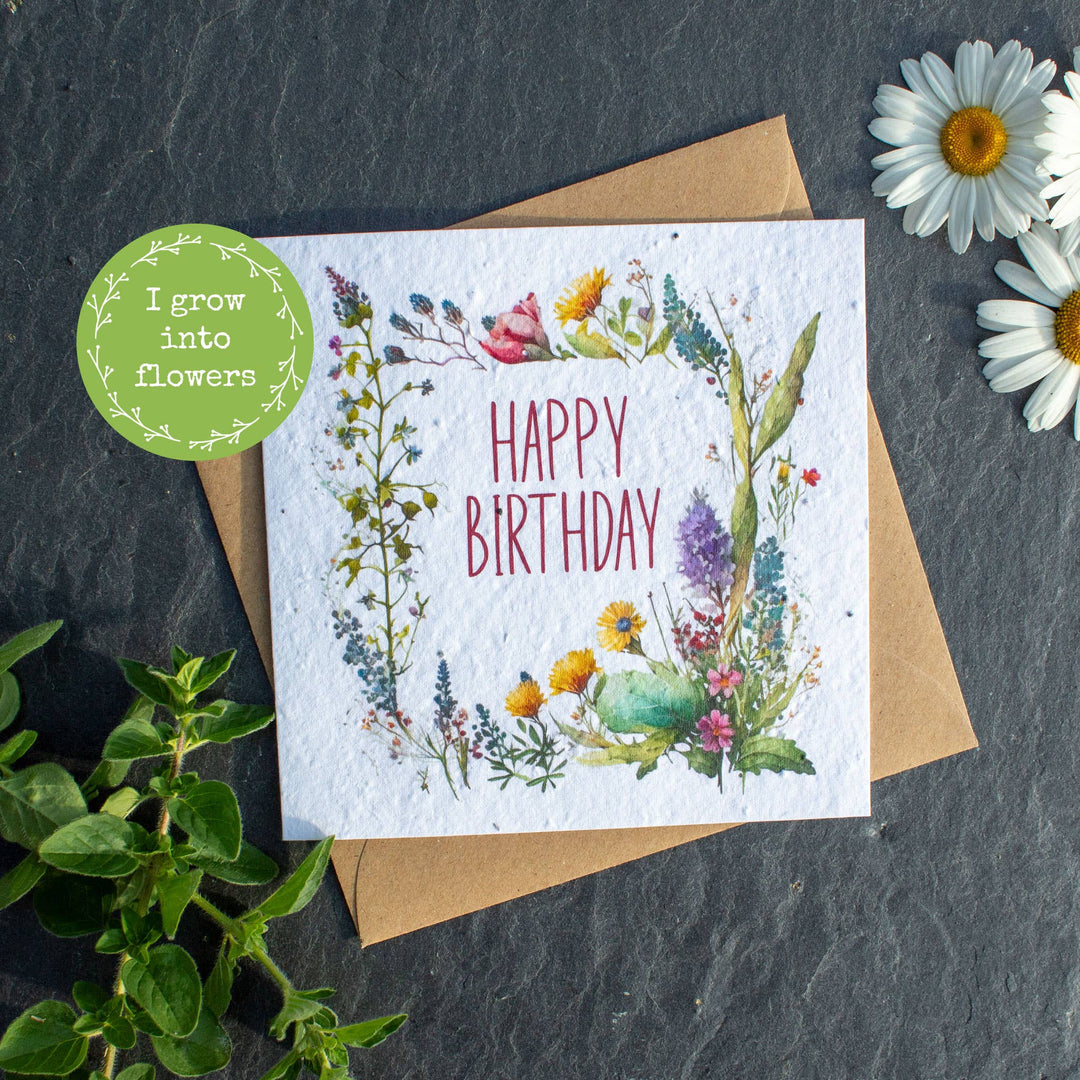 A square, plantable birthday card with 'Happy Birthday' written on it, adorned with a colorful flower wreath, placed on a grey background with a recycled brown kraft paper envelope and a daisy flower behind it.