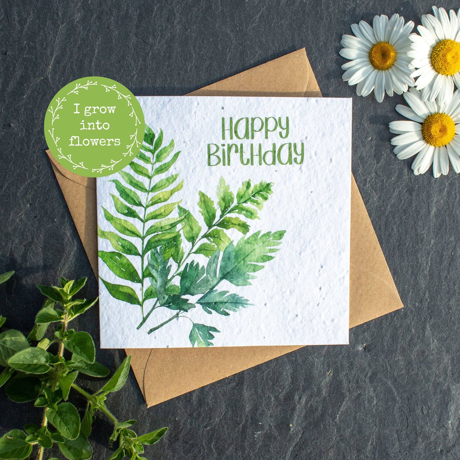 Birthday card with greenery design, envelope, and daisies on a dark surface