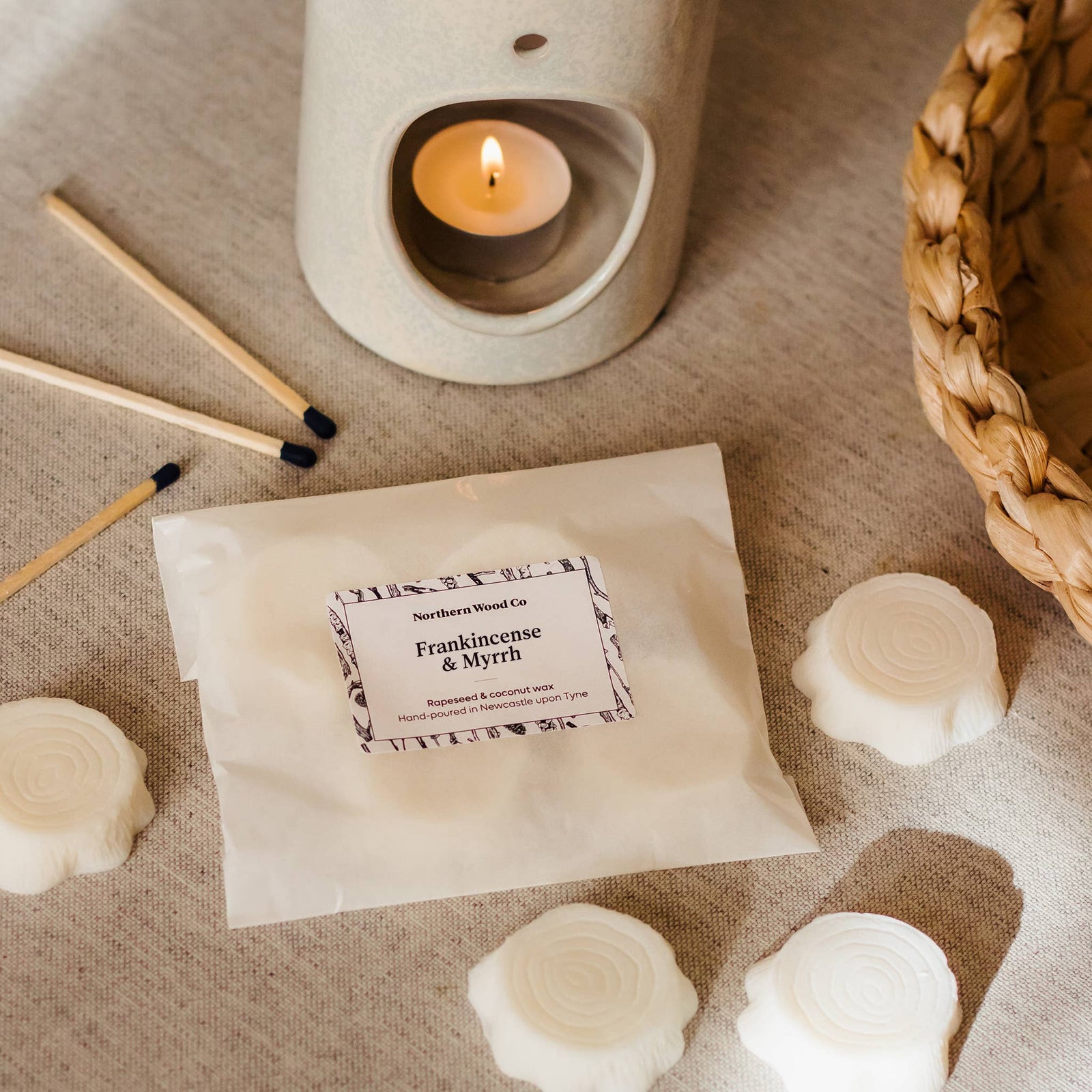  Wax melts shaped like small tree stumps, with a burning candle in the background, indicating use. Packaging is visible with branding, and the scene is set on a textured fabric surface.