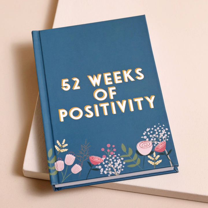 Write down your thoughts, feelings and reflections on each day of the year, this diary focuses on the positive. The pages are encased in a teal cover adorned with illustrations of pink flowers and botanical details.
