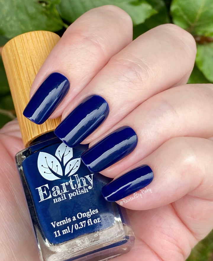 Hand with blue nail polish holding an Earthy nail polish bottle against a green leafy background