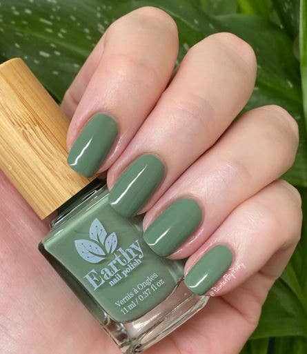 Hand with green nail polish holding an Earthy Nail Polishes bottle against a leafy background