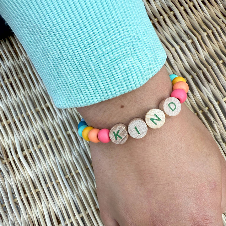 Colourful beaded bracelet spelling 'KIND' on a person's wrist against a woven background.