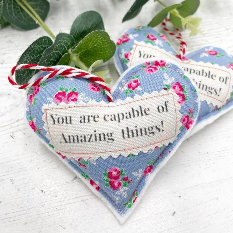 Two heart-shaped decorations made of cotton and felt, with blue floral fabric and the phrase 'You are capable of Amazing things!' in the center, tied with twine.
