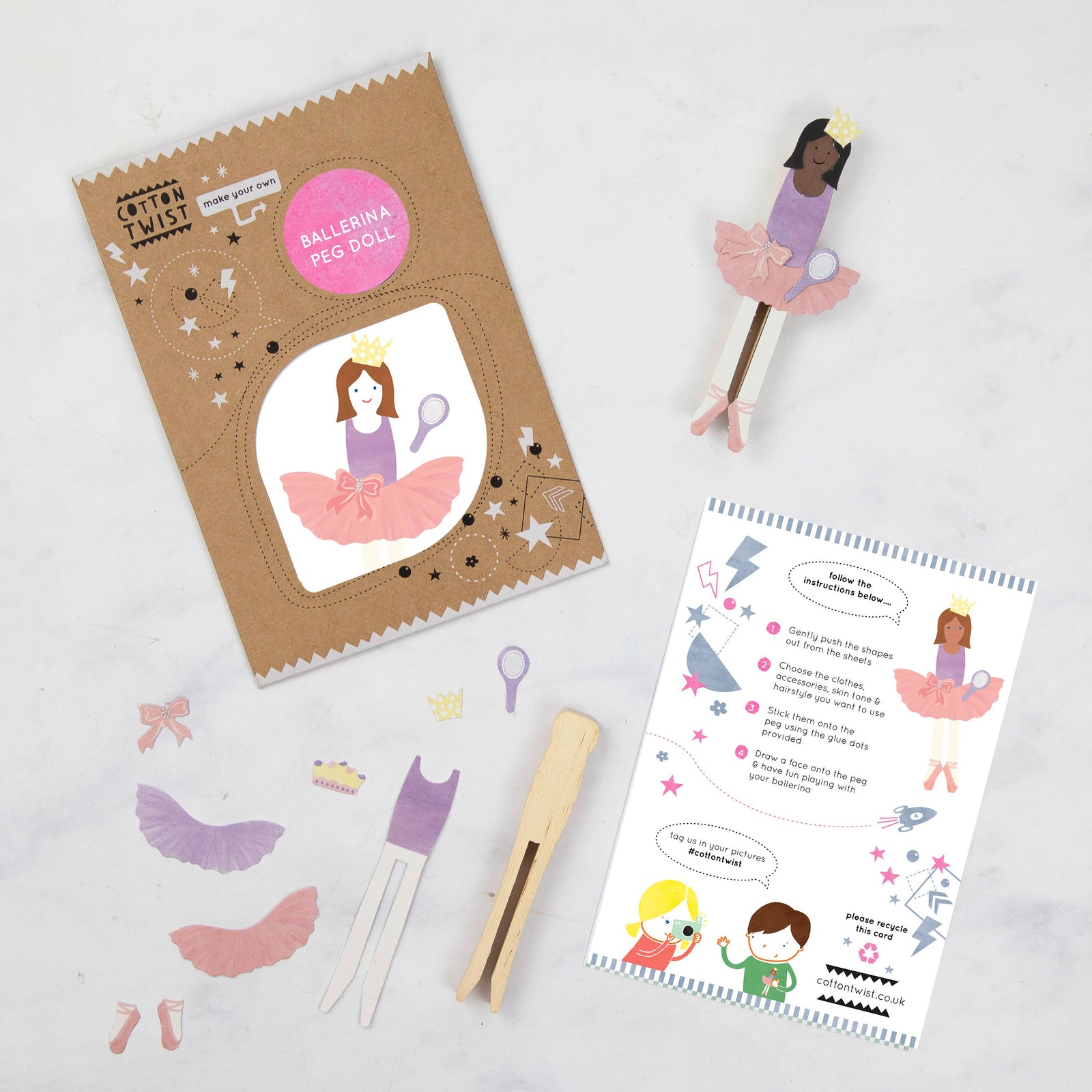 The image shows a ballerina peg doll kit, including a cardboard cut-out of a ballerina in a pink tutu, a wooden peg doll, and various accessories such as a mirror, crown, bow, bodysuit, and ballet shoes. There is also a printed instruction card.