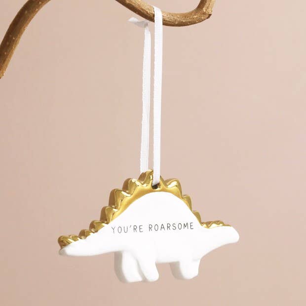 A white ceramic dinosaur-shaped hanging decoration with a golden ribbon and the text 'you're roarsome' printed on its back.